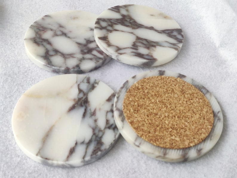 Pu&ograve; includere: Set di quattro sottobicchieri rotondi in marmo con un motivo a marmo bianco e grigio. Un sottobicchiere ha un retro in sughero.