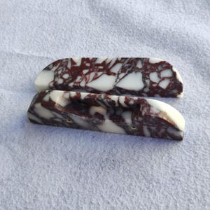 Op de afbeelding: Twee decoratieve kastgrepen met een uniek marmerpatroon. De grepen zijn wit met bordeauxkleurige adering en hebben een gebogen vorm. Elke greep heeft een klein, goudkleurig accent.