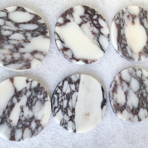 Pu&ograve; includere: Sei sottobicchieri rotondi in marmo bianco con un motivo a spirale di venature grigie e viola. I sottobicchieri sono disposti in due file da tre.