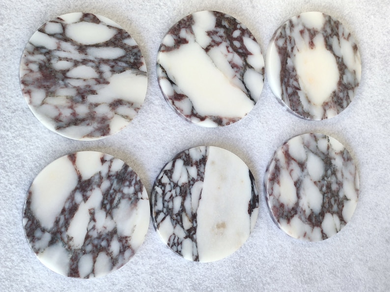 Pu&ograve; includere: Sei sottobicchieri rotondi in marmo bianco con un motivo a spirale di venature grigie e viola. I sottobicchieri sono disposti in due file da tre.