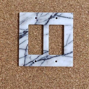 Puede incluir: Una placa de cubierta de interruptor de luz doble de mármol blanco con vetas negras. La cubierta tiene dos aberturas rectangulares y cuatro orificios para tornillos. El diseño de mármol añade un toque de elegancia a cualquier habitación.