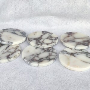 Pu&ograve; includere: Sei sottobicchieri rotondi in marmo bianco con venature grigie. I sottobicchieri sono disposti a semicerchio.