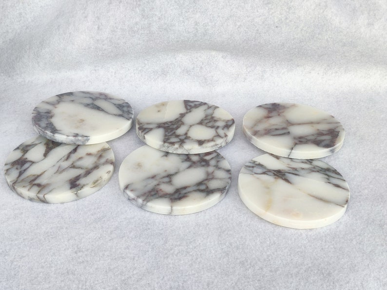 Pu&ograve; includere: Sei sottobicchieri rotondi in marmo bianco con venature grigie. I sottobicchieri sono disposti a semicerchio.