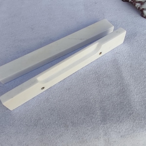 Puede incluir: Dos piezas de mármol blanco. Una es un bloque rectangular, la otra es una pieza rectangular con una hendidura curva y dos pequeños detalles metálicos. Las piezas están sobre una superficie blanca.