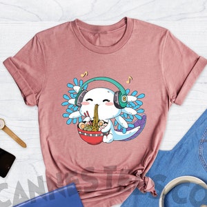 May include: A heather mauve t-shirt with a cartoon axolotl wearing headphones and eating ramen noodles. The text "Cannes Tees Co" is on the top left of the image.