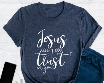 May include: A heather navy blue t-shirt with the text "Jesus I trust in you" in white lettering. The shirt is displayed on a wooden background with other items.