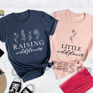 K&ouml;nnte beinhalten: Zwei T-Shirts mit einem floralen Design und Text. Das blaue Shirt sagt "Raising Wildflowers" und das rosa Shirt sagt "Little Wildflower".