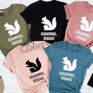 May include: Five t-shirts with different color backgrounds and a white squirrel graphic with the text "Squirrel Squad" on each shirt. The shirts are in the colors heather olive, black, heather mauve, heather peach, and heather deep teal.