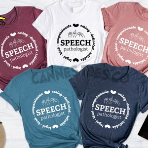 Op de afbeelding: Vijf verschillende t-shirts met een wit cirkelvormig ontwerp met de tekst "SPEECH pathologist" en een handgebaar voor het woord "speech". De kleuren van de t-shirts zijn heidebruin, wit, heidepaars, diep teal en heideblauw.