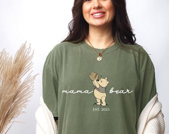 May include: A green t-shirt with a cartoon Winnie the Pooh bear design. The text "mama bear est. 2025" is printed on the shirt.