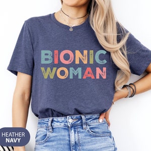 May include: A heather navy blue t-shirt with the text "Bionic Woman" in colorful, distressed lettering.