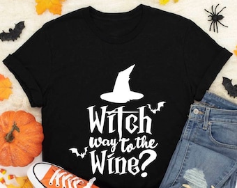 May include: Black t-shirt with the text "Witch way to the Wine?" in white, featuring a witch hat and bats. The shirt is styled with orange sneakers, blue jeans, and Halloween decorations like a pumpkin and a skull.