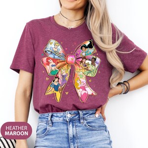 May include: A heather maroon t-shirt featuring a colourful bow design with various Disney princesses. The bow is made up of different princess characters, including Cinderella, Ariel, Belle, and Aurora.