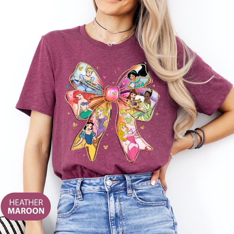 May include: A heather maroon t-shirt featuring a colourful bow design with various Disney princesses. The bow is made up of different princess characters, including Cinderella, Ariel, Belle, and Aurora.