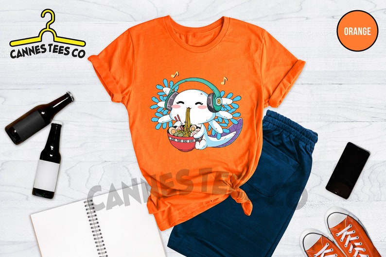 May include: Orange t-shirt with a cartoon axolotl wearing headphones and eating ramen noodles. The text "Cannes Tees Co" is on the top left corner of the image. The word "ORANGE" is in a circle on the top right corner of the image.