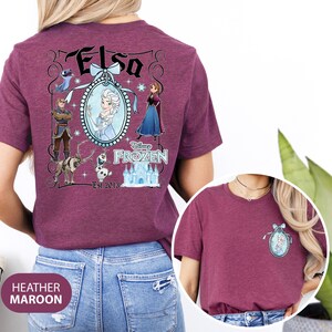 May include: Heather maroon t-shirt featuring a Disney Frozen design. The design includes the name "Elsa" in a decorative font, an image of Elsa, and other characters from the movie. The shirt also has a smaller design on the front.