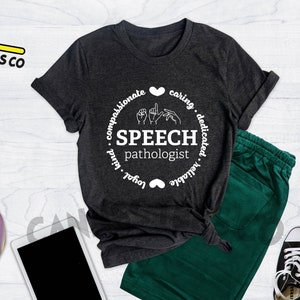 Op de afbeelding: Heather donkergrijs T-shirt met een wit cirkelontwerp met de tekst "SPEECH pathologist" en gebarentaal voor het woord "speech". De cirkel is omgeven door de woorden "compassionate, caring, dedicated, reliable, loyal, kind".