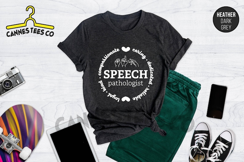 Op de afbeelding: Heather donkergrijs T-shirt met een wit cirkelontwerp met de tekst "SPEECH pathologist" en gebarentaal voor het woord "speech". De cirkel is omgeven door de woorden "compassionate, caring, dedicated, reliable, loyal, kind".