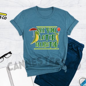 May include: A teal heather t-shirt with a graphic of two alligators, one holding an umbrella and the other holding a balloon. The text "See you later alligator" is printed on the shirt.
