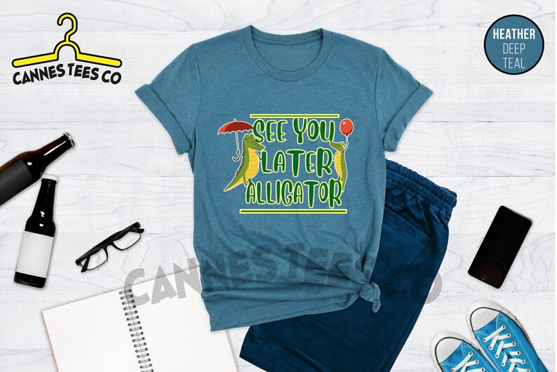 May include: A teal heather t-shirt with a graphic of two alligators, one holding an umbrella and the other holding a balloon. The text "See you later alligator" is printed on the shirt.