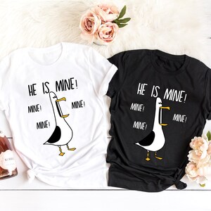 May include: Two white t-shirts with black text and a cartoon seagull graphic. The text on the shirts reads "He is mine!" with the word "mine" repeated three times.