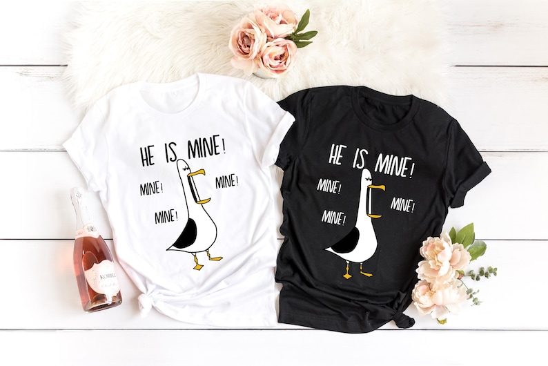 May include: Two white t-shirts with black text and a cartoon seagull graphic. The text on the shirts reads "He is mine!" with the word "mine" repeated three times.