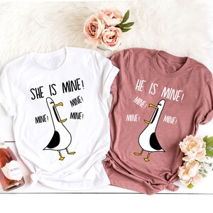 May include: Two white t-shirts with a cartoon seagull graphic and the text "She is mine!" and "He is mine!" with the word "mine" repeated multiple times. The shirts are on a white surface with a pink and white floral arrangement.