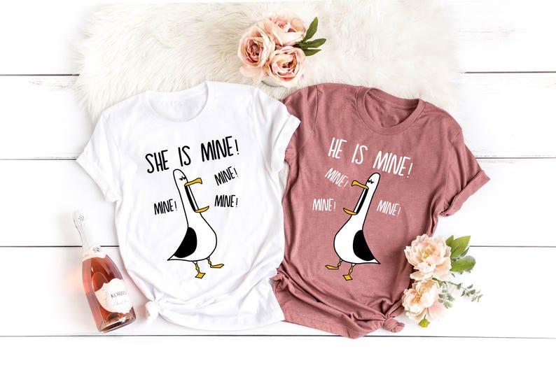 May include: Two white t-shirts with a cartoon seagull graphic and the text "She is mine!" and "He is mine!" with the word "mine" repeated multiple times. The shirts are on a white surface with a pink and white floral arrangement.