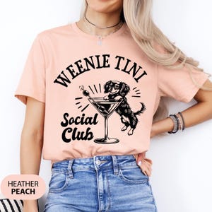 May include: A peach colored t-shirt with black text that reads "Weenie Tini Social Club". A black and white illustration of a dachshund holding a martini glass is above the text.