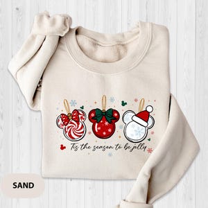 May include: Sand-colored sweatshirt featuring a festive graphic of three Mickey Mouse-shaped ornaments. The ornaments are red and white with candy cane, bow, and snowflake designs. The text "Tis the season to be jolly" is printed below the ornaments.