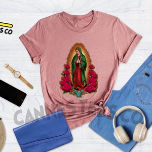 May include: Heather mauve t-shirt with a graphic of Our Lady of Guadalupe, a religious figure, surrounded by red roses. The t-shirt is laid flat on a white wooden surface with other accessories.