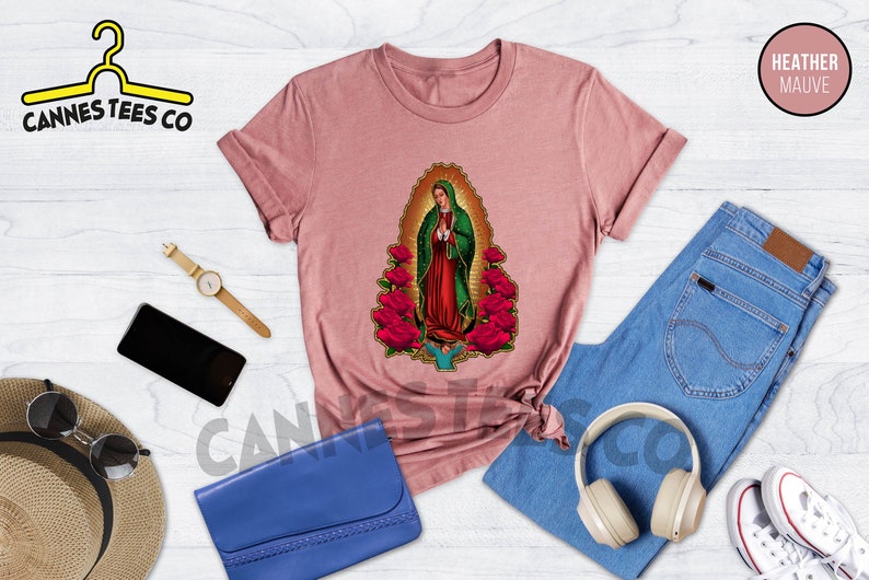 May include: Heather mauve t-shirt with a graphic of Our Lady of Guadalupe, a religious figure, surrounded by red roses. The t-shirt is laid flat on a white wooden surface with other accessories.