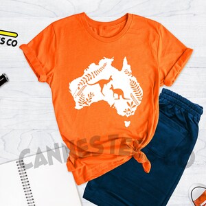 Może przedstawiać: Pomarańczowy T-shirt z białym siluetem kontynentu australijskiego z dwoma kangurami i roślinnością. Koszulka leży płasko na drewnianej powierzchni z niebieskimi spodenkami, notesem, dwoma butelkami piwa, telefonem kom&oacute;rkowym i dwiema parami czerwono-białych but&oacute;w sportowych.