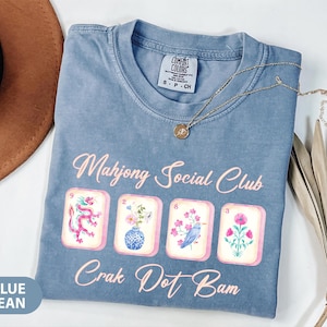 May include: A blue jean colored t-shirt featuring the text Mahjong Social Club and Crak Dot Bam in light pink. Below the top text are four mahjong tiles displaying a pink dragon, a vase with flowers, a bird on a branch, and a pink flower.