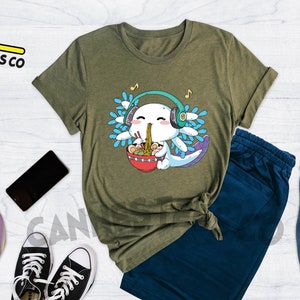 May include: A green t-shirt with a cartoon axolotl wearing headphones and eating ramen. The axolotl is surrounded by blue and green leaves. The text "Cannes Tees Co" is in the top left corner of the image. The text "HEATHER OLIVE" is in the top right corner of the image.
