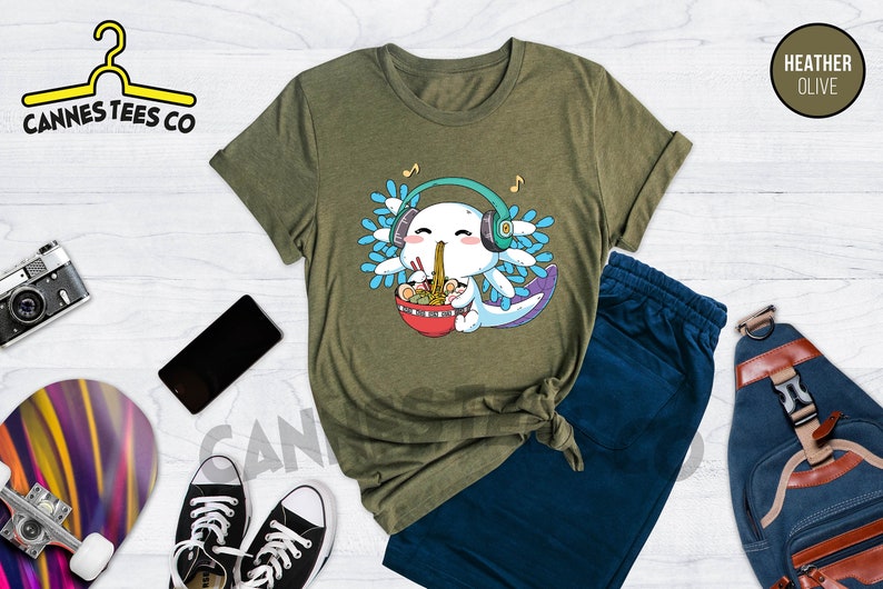 May include: A green t-shirt with a cartoon axolotl wearing headphones and eating ramen. The axolotl is surrounded by blue and green leaves. The text "Cannes Tees Co" is in the top left corner of the image. The text "HEATHER OLIVE" is in the top right corner of the image.