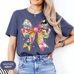 May include: A heather navy blue t-shirt featuring a large colourful bow design with Disney princesses. The bow is made up of different princesses, including Cinderella, Ariel, Belle, and Snow White.