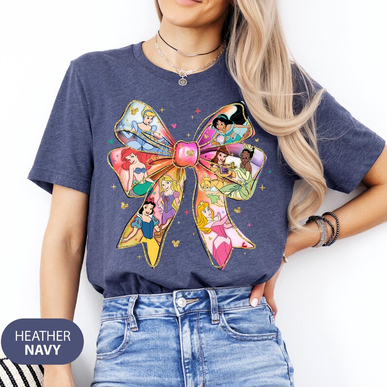 May include: A heather navy blue t-shirt featuring a large colourful bow design with Disney princesses. The bow is made up of different princesses, including Cinderella, Ariel, Belle, and Snow White.