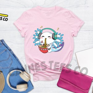 May include: Pink t-shirt with a cartoon axolotl wearing headphones and eating ramen noodles. The axolotl has blue and green gills and is smiling. The text "Cannes Tees Co" is on the top left of the image.