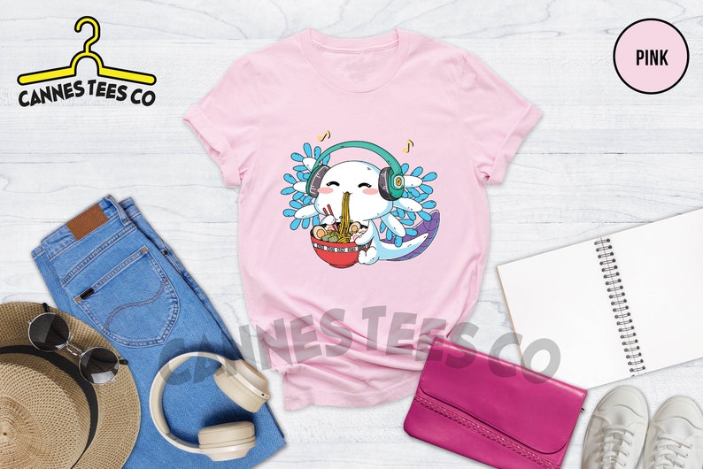 May include: Pink t-shirt with a cartoon axolotl wearing headphones and eating ramen noodles. The axolotl has blue and green gills and is smiling. The text "Cannes Tees Co" is on the top left of the image.