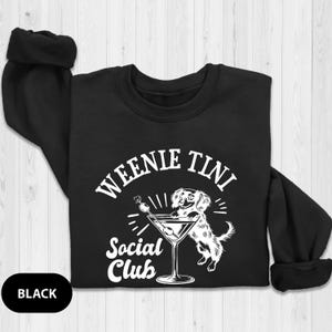 May include: Black sweatshirt with a white graphic of a dachshund holding a martini glass. The text on the graphic reads "Weenie Tini Social Club".