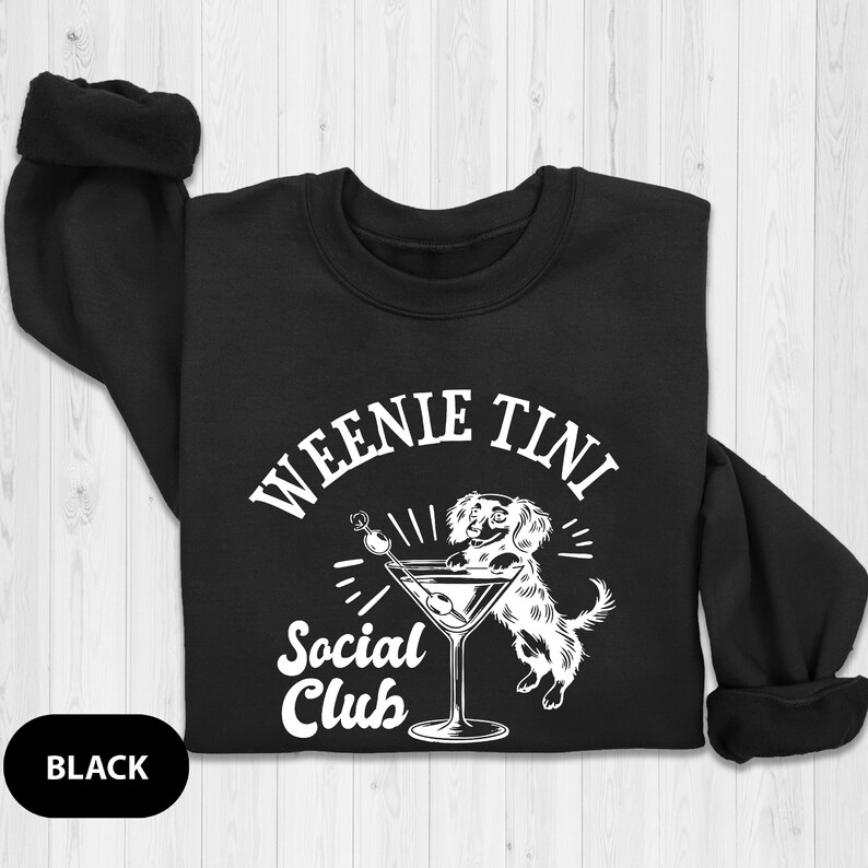 May include: Black sweatshirt with a white graphic of a dachshund holding a martini glass. The text on the graphic reads "Weenie Tini Social Club".