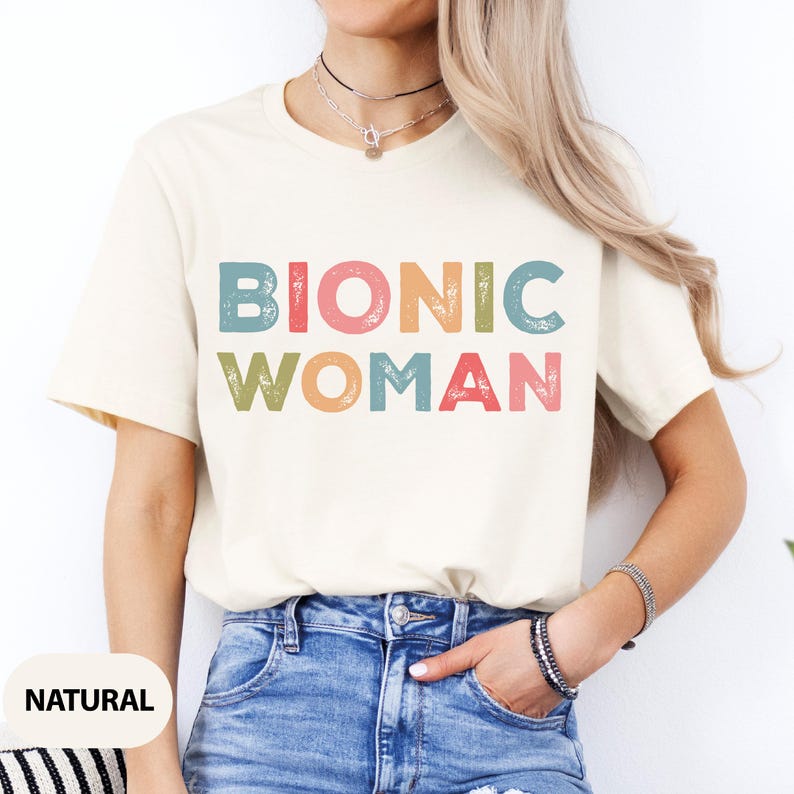 May include: A white t-shirt with the text "Bionic Woman" in colorful, distressed lettering. The shirt is being worn by a person with long blonde hair and is paired with blue jeans.