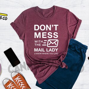 May include: Heather maroon t-shirt with the text "Don't Mess With The Mail Lady (I Know Where You Live)" in white. The shirt features a stylized white envelope graphic.