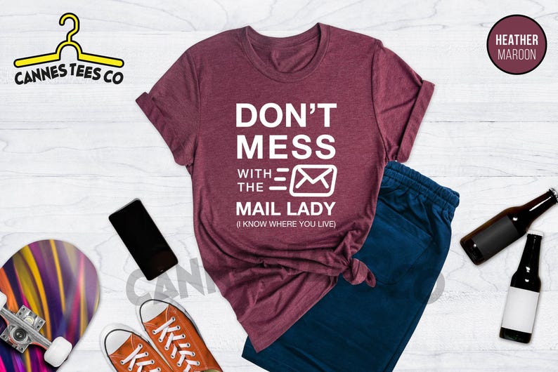 May include: Heather maroon t-shirt with the text "Don't Mess With The Mail Lady (I Know Where You Live)" in white. The shirt features a stylized white envelope graphic.