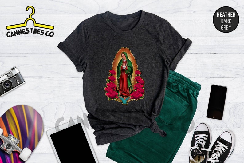 May include: A dark heather grey t-shirt with a graphic of the Virgin Mary surrounded by red roses. The text "Cannes Tees Co" is visible in the top left corner of the image.