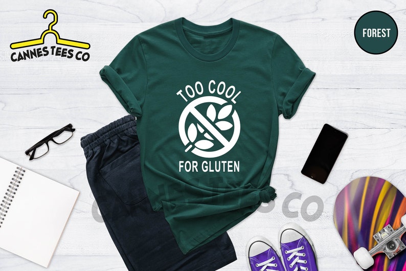 May include: A green t-shirt with a white graphic that says "Too Cool For Gluten" and a crossed-out wheat symbol. The shirt is laid out on a white wooden surface with a pair of black eyeglasses, a notebook, a pair of purple sneakers, a black smartphone, and a colorful skateboard.
