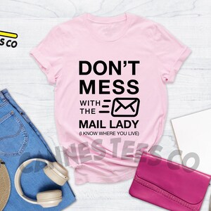 May include: Pink t-shirt with black text that reads "Don't Mess With The Mail Lady (I Know Where You Live)" and a black envelope graphic.