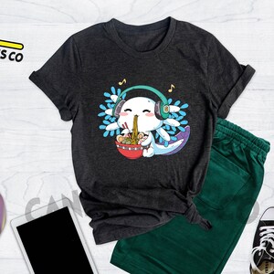 May include: A dark heather grey t-shirt featuring a cartoon axolotl wearing headphones and eating ramen noodles. The axolotl is blue and white with a pink nose and mouth. The ramen noodles are in a red bowl with chopsticks. The text "Cannes Tees Co" is on a yellow hanger in the top left corner of the image.