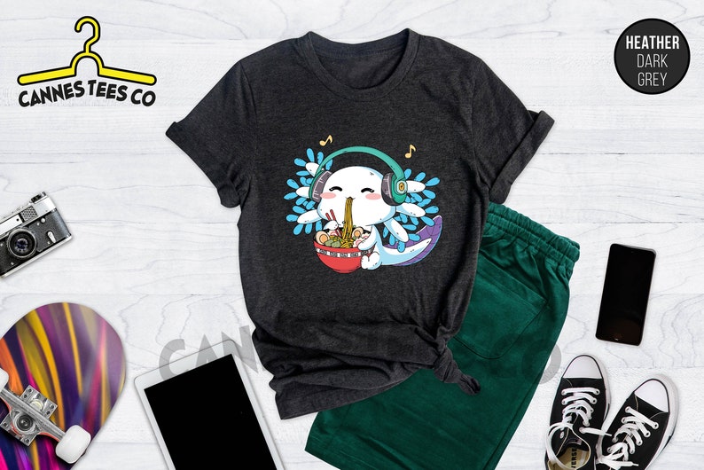 May include: A dark heather grey t-shirt featuring a cartoon axolotl wearing headphones and eating ramen noodles. The axolotl is blue and white with a pink nose and mouth. The ramen noodles are in a red bowl with chopsticks. The text "Cannes Tees Co" is on a yellow hanger in the top left corner of the image.